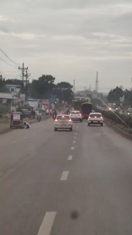 The video shows a view from a vehicle moving down a road. The sky is overcast. Several cars are driving on the road, including a white car in the foreground, a white car in the middle distance, and a white car behind it. A large yellow truck is also visible further down the road. On the left side of the road, there are shops and buildings, with power lines and poles. There are also people on motorcycles and walking on the side of the road. The road has white dashed lines marking the lanes.