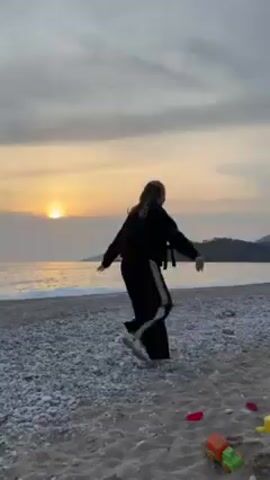 A person stands on a beach at sunset, with their back to the camera. They are wearing a black jacket and pants with a white stripe down the side, and a dark backpack. The person has blonde hair pulled back from their face.  They spread their arms out and then dance, making gestures with their hands. The beach is composed of light gray pebbles, with some patches of sand near the bottom of the frame. There are a few scattered colorful plastic toys on the sand. The water is a calm, dark gray and the sun is a bright orange disk setting on the horizon. A tree-covered land mass is visible in the distance. The sky is mostly cloudy with light orange hues near the setting sun.