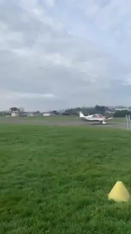 A small airplane takes off from a runway at an airfield on a partly cloudy day.  The airplane is white with red trim and is taking off toward a distant town. A film crew stands next to a white van near the runway. They appear to be filming the airplane as it takes off. The airfield is surrounded by green grass and a paved area. There are yellow cones placed on the grass.