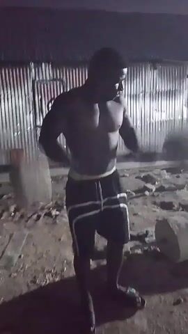 A dark-skinned male is seen performing weightlifting exercises in an outdoor, dimly lit environment. He is shirtless and wearing dark shorts with a white stripe. The male is holding a barbell with weights on each end, performing a row exercise, bending over at the waist. The background appears to be an open area with some structures, possibly a corrugated metal fence and some debris on the ground. The lighting suggests it is nighttime, with some artificial light sources casting shadows.