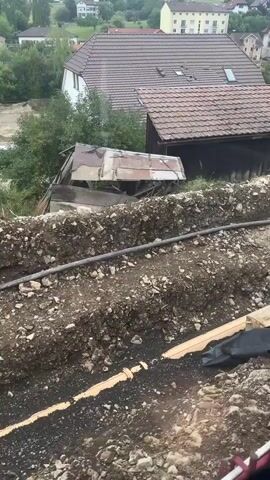 This is a downward-angle video taken from above a construction site. In the foreground, a black plastic sheeting covers a sloped area of dirt and gravel. A yellow and black compactor machine is visible on the sheeting. Behind this, a trench has been dug into the side of a hill. The trench is filled with dirt and rocks, and a dark pipe runs through it. Beyond the trench, a building with a brown tiled roof is visible. Further in the background, houses and trees are seen on a hillside. The sky appears overcast.