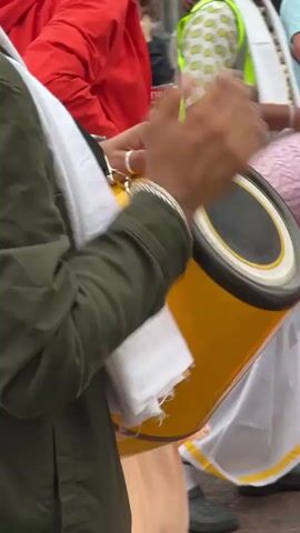 The video shows a close-up of a person playing a yellow drum with their hands. The drum is cylindrical and has black rims. The person is wearing a green jacket, a white cloth draped around their neck, and light-colored pants. They are also wearing bracelets on their wrist. In the background, other people are visible, some of whom are also holding drums. One person in the background is wearing a red jacket and another is wearing a pink shirt. The setting appears to be outdoors, possibly a street or parade, with a crowd of people. The overall impression is of a musical performance or celebration.