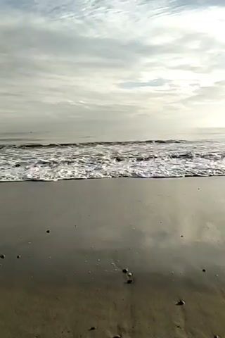The video depicts a beach scene on a cloudy day. The camera is positioned close to the ground, capturing the wet sand and the foamy waves rolling in. The sky is overcast with varied cloud formations, creating a somewhat diffused light. There are small pebbles scattered on the beach. The horizon is visible in the distance, blurring into the sky. The video is still, with only the gentle movement of the waves.