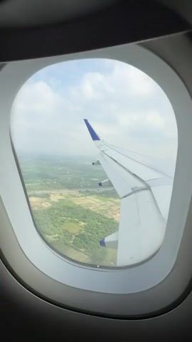 The video shows a view from an airplane window. Outside the window, the wing of the airplane is visible, along with a landscape of green fields, trees, and some buildings. The sky is cloudy. The camera is static and the view outside the window is also static, indicating the airplane is in flight.