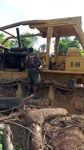 A man wearing a dark vest with "JAMES 6" on the back and shorts stands on the tracks of a large, yellow bulldozer, which is partially covered in mud. The bulldozer has "D-8K" visible on its side, suggesting it's a Caterpillar D-8K model. The man is positioned near the engine compartment, appearing to be working on or inspecting the heavy machinery. The scene is set outdoors in a rural environment under a clear, bright sky, likely during the day. The ground is reddish-brown earth, indicative of a construction or land-clearing site, with exposed roots and scattered green vegetation. In a later shot, the bulldozer's blade is seen actively moving earth, carving out a dirt road. Another person walks along this newly formed path in the distance, surrounded by trees. The activity suggests land development or road construction in Otunade, Nigeria.