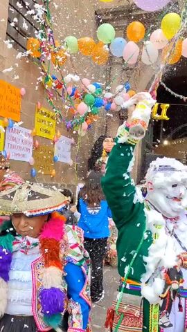The video shows a group of people celebrating outdoors. They are standing in front of a stone building decorated with balloons and the word "COMADRES" written in gold letters. Some people are dressed in colorful traditional outfits. One person sprays foam from a can into the air, covering themselves and others. Confetti is also raining down. The atmosphere appears festive and joyful.