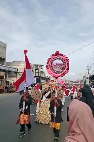 A group of people are participating in a parade. In the foreground, two individuals in black shirts and gold skirts hold red and white poles. Behind them, a person wears an elaborate costume with gold and brown feather-like embellishments and a gold crown, holding a sign. A large, circular red decoration with a picture of a building is held aloft. The parade is taking place on a paved street lined with buildings and a few vehicles. Other participants in red and white attire are visible further down the street. The sky is overcast.