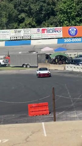 A white Nissan 350Z with a white and red front bumper is shown drifting on a race track. In the background, there are several tents and trailers. There are also advertisements on a wall behind the track. Another silver Nissan 350Z is also visible on the track. The area appears to be a race track with spectators in the background. The weather appears to be sunny and warm.