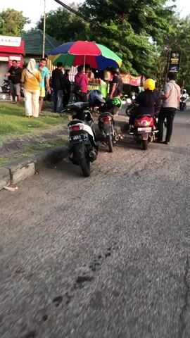 The video shows an outdoor scene with several motorcycles parked on a paved surface. A colorful umbrella is set up, likely for a vendor. Several people are gathered around the umbrella, some on motorcycles, some standing. The environment appears to be a roadside or market area with some greenery and trees in the background. The lighting suggests it is daytime, possibly late afternoon or early evening.