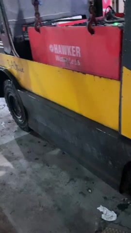 The video shows a forklift in a warehouse setting. The forklift is primarily yellow with black accents and the words "T2Active" visible on the side. A large, red, rectangular Hawker Waterless battery is being lifted out of the forklift using an overhead crane system. The crane system has yellow components, purple straps, and metal hooks. The lifting device is labeled with "WLL 3.200 KG," indicating its working load limit. The background shows blurred stacks of goods, suggesting the interior of a busy warehouse. The floor appears wet and possibly oily. There is a forklift with the driver visible in the background.
