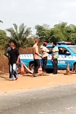 The video shows a roadside scene with a blue and white pickup truck with what appears to be emergency lights on top, parked near some orange traffic cones. Several people are standing near the truck, including at least one individual wearing a red cap. A motorcycle passes by in the foreground, partially obscuring the view. The background includes trees and other vegetation. The road appears to be paved but somewhat worn.