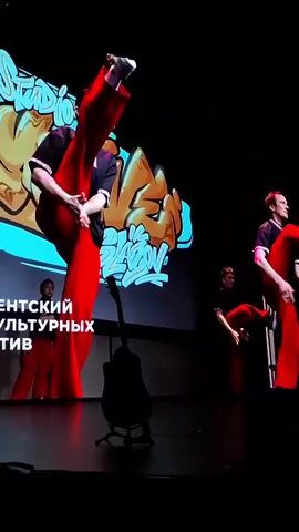 A recording depicts a stage performance. A man in a black top and red pants is dancing on a stage with a graffiti-style backdrop. There's a silhouette of an upright bass near him. The video transitions to a view of an audience, many raising their hands. Later, a woman in a grey sweater and jeans is seen in the audience. The video returns to the stage, now showing a presentation featuring individuals seated on chairs behind a bass silhouette.