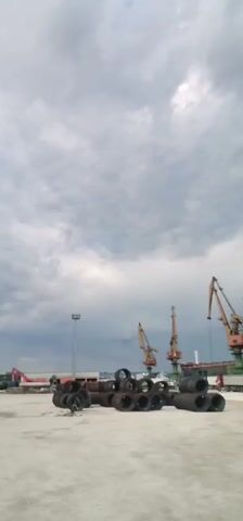 The video depicts a dockyard or port setting under a cloudy sky. Stacks of what appear to be metal pipes or coils are in the foreground. Behind them are several large cranes, seemingly for loading and unloading cargo. In the background, a large cargo ship is docked alongside the pier. Several trucks are parked next to the ship, likely involved in the transportation of goods. There are a couple of birds flying across the sky. A single person is briefly visible walking on the pier.
