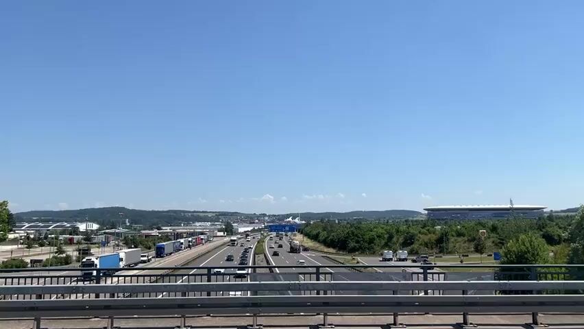 This is a static wide shot of a highway with traffic under a clear blue sky. In the foreground, there is a metal railing. The highway has multiple lanes and is filled with cars and trucks. To the left of the highway, there is a rest stop area with many parked trucks. In the distance, there are trees, hills, and a large modern building with a distinctive roof structure. The sun is shining brightly, indicating it is daytime. The video appears to be filmed from an elevated position, likely a bridge or overpass. There are no people visible in the video.
