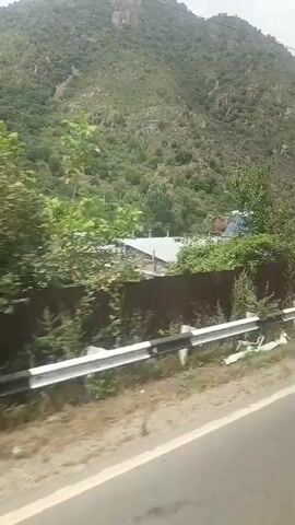 The video appears to be shot from a moving vehicle. It shows a mountainous landscape with lush green vegetation covering the slopes. In the foreground, there is a road with a metal guardrail. Further down the road, several structures are visible, including a large shed with open doors, a building with multiple windows and a satellite dish, and a green truck filled with materials. There are also piles of bricks and coiled pipes. The sky is overcast.