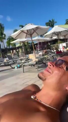 A low-angle shot shows a man relaxing in a lounge chair outdoors on a sunny day. He is shirtless, wearing sunglasses and a beaded necklace. The background consists of palm trees, white umbrellas, lounge chairs, and a patio area. As the video continues, the camera angle adjusts, revealing a view of the ocean behind a railing. The sky is bright blue with scattered white clouds.