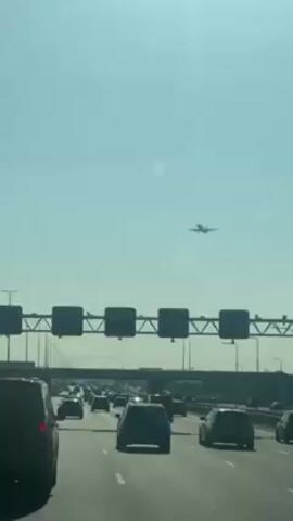 The video shows a highway with heavy traffic. The camera is likely inside of a vehicle. An airplane is seen in the sky above the highway. The sky is light blue and there are lamp posts along the highway. There are metal sign gantries above the highway.