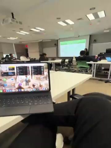 The video shows a classroom setting, likely a university or college lecture hall. Several students are seated at desks, some with laptops open, listening to a lecture. A projector displays a presentation on a screen at the front of the room. The camera is focused on an iPad with a keyboard case sitting on a desk in the foreground. The iPad screen is visible, displaying various icons and widgets. Overall, the scene depicts a typical classroom environment during a lecture.