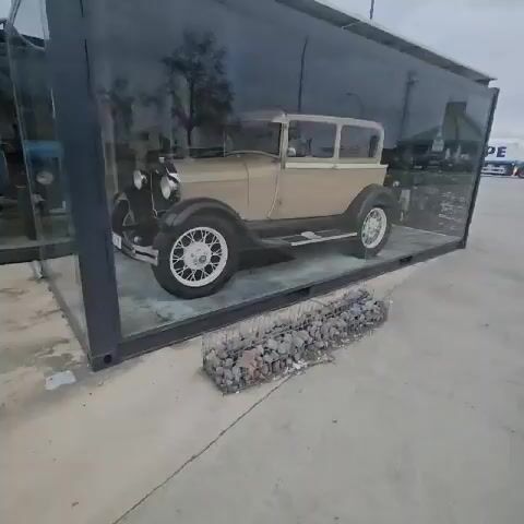 The video shows two vintage cars displayed in glass enclosures. The first car is a tan and black Ford with the license plate "Z-8699." The second car is blue and black and has a scale in front of it. Both cars are displayed on concrete. Outside the enclosures, there are stone-filled gabion baskets. The sky is overcast.