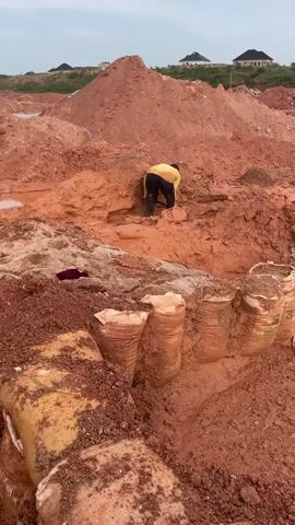 This media file depicts an outdoor scene at a construction or excavation site in Rayfield, Nigeria. The primary subject is a person wearing a yellow shirt and dark pants, actively engaged in digging or excavating a trench in the reddish-brown soil. The ground is uneven, with large mounds of dirt and several partially filled sandbags lining a trench in the foreground. These sandbags appear to be used for erosion control or to shore up the excavation.

The setting suggests a significant earth-moving operation is underway. In the background, more large piles of red soil are visible, indicative of extensive digging. Further in the distance, several houses with dark roofs are situated on a hill, suggesting a residential area nearby. The sky is overcast, hinting at a cloudy day or possibly early morning or late afternoon, though the overall lighting does not strongly indicate a specific time of day. No visible text is present in the media. The activity appears to be manual labor, with the individual focused on their task in a somewhat muddy and disturbed environment. The mood conveyed is one of diligent work amidst a large-scale construction project.