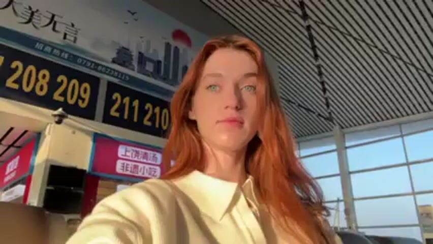 A young woman with red hair and pale blue eyes is facing the camera inside what appears to be an airport terminal. She is wearing a beige collared shirt. Behind her is a sign with Chinese characters and a series of numbers (208, 209, 211, 210). There's also a smaller red sign with Chinese writing underneath the numbered sign. The ceiling is white with a linear, possibly slatted design. Light is entering the scene from the right, illuminating the woman’s face. The camera angle is slightly below eye level.
