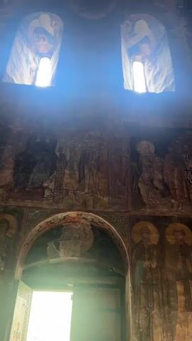 The video pans across the interior of a dimly lit, ancient stone structure, likely a church or monastery, with frescoes adorning the walls. Sunlight streams in from two high windows, creating beams of light that illuminate dust motes in the air. The frescoes depict religious figures in a somewhat faded, archaic style. There is an arched doorway in the foreground with light shining through it, indicating an exit or entrance. Several people, their faces mostly obscured or out of focus, are gathered in the lower part of the frame, some appearing to be observing the frescoes or interacting near the doorway. The stone architecture shows signs of age and wear, with some crumbling sections visible. The overall atmosphere is one of historical reverence and solemnity.