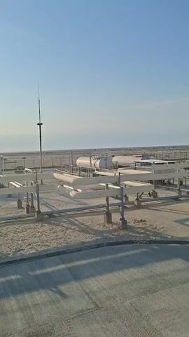 The video shows an outdoor industrial facility on a clear, sunny day. The foreground features a concrete area with shadows cast by an unseen structure. Behind this, a sandy area is dotted with concrete supports and a network of white pipes running horizontally at a moderate height. To the left, a tall, thin lightning rod with a pointed top stands vertically. In the background, several large white cylindrical tanks are visible, along with more fencing and flat, arid terrain extending to the horizon. The sky is a pale blue with no clouds.