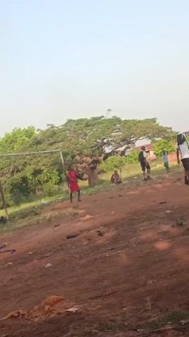 The video shows several young people playing a sport, possibly soccer, on a dirt field. The field is mostly dirt with some grass patches. In the background, there is a building, possibly a house or school, and trees. The sky is clear with a light blue hue. The video follows the trajectory of the ball as it is kicked into the air. The people are dressed casually and appear to be enjoying themselves.