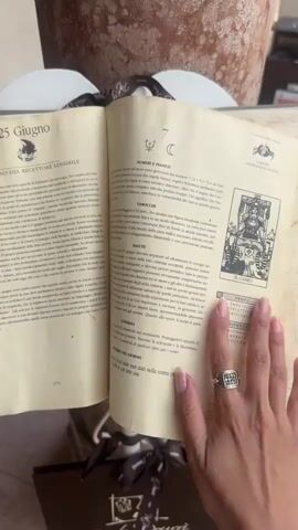A person's hand with manicured nails and rings turns the pages of an old, worn book with yellowed pages. The book appears to be a daily horoscope or astrological guide, with dates and text in Italian. One page visible has the date "25 Giugno" (June 25th) and the heading "IL GIORNO DEL RICETTORE SENSIBILE" (The Day of the Sensitive Receiver). Another page features a tarot card illustration. The background shows a large, textured pot and hints of other objects. The lighting is natural, suggesting daytime.