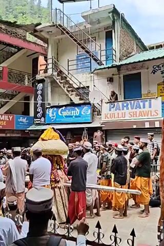 A vibrant religious procession is underway in a narrow street or town square, likely in a mountainous region given the green hillside in the background. A group of men, many wearing traditional Himachali caps (Kullu caps), are carrying a palanquin or ceremonial chariot adorned with yellow and red fabric. The palanquin appears to hold a deity or religious symbol, and its carriers are dressed in traditional dhotis or similar lower garments, some in orange/yellow and others in red.

The setting is characterized by multi-story buildings with balconies and external staircases, indicative of a busy town or village. Several shops line the street. A notable detail is a man on an upper-level balcony, also wearing a Himachali cap, observing the procession and holding a long stick or rod. A satellite dish is mounted on the building beside him.

Visible text on shop signs includes "PARVATI KULLU SHAWLS" and "GOLIND" (partially visible). Another sign above a blue door reads "WELCOME." The time of day appears to be daytime, with clear weather. The scene exudes a sense of cultural celebration and community engagement, typical of religious festivals in the Himachal Pradesh region.