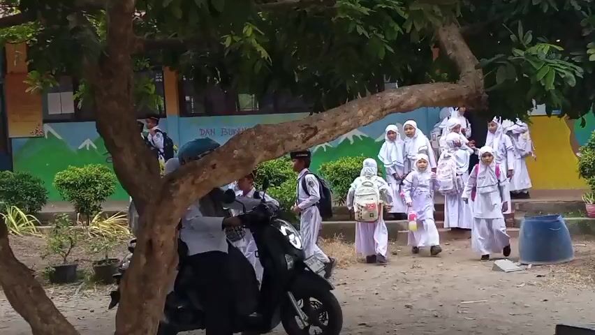The video shows a group of children, mostly wearing white uniforms and hijabs, walking in an outdoor area with a building in the background. Some children are carrying backpacks. A motorcycle with two people on it is visible in the foreground, partially obscured by a tree. The building in the background has colorful murals on its exterior walls, depicting mountains and trees. The ground is dusty and appears to be a courtyard or playground. The overall impression is of a school environment.