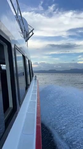 A video taken from the side of a white boat with a red stripe along its hull, moving across the water. The boat creates a significant white, foamy wake that streams behind it. The camera is positioned looking out from the boat's side, showing several dark-tinted windows reflecting the sky. In the distance, a serene expanse of water meets a distant shoreline with silhouetted mountains or hills under a partly cloudy sky. The clouds are soft, with hints of warm light, suggesting late afternoon or early evening. The scene depicts travel by sea, likely around Jungutbatu, Indonesia, as indicated by the context. No people or visible text are present.