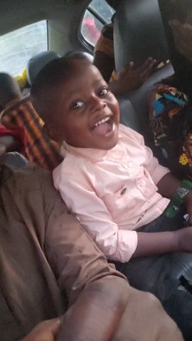 A young, dark-skinned child, likely a boy, is seen laughing and smiling joyfully inside a vehicle. He is wearing a light pink long-sleeved collared shirt and brown pants. His short, dark hair frames a face lit up with a wide, toothy grin. A green bracelet adorns his left wrist, and he holds a small, light-colored object in his left hand. The child is positioned lying down, possibly on an adult's lap or across seats, with his head tilted upwards towards the camera.

The interior of the vehicle, possibly a car or van, features grey upholstery. Parts of other individuals are visible: an adult's arm with patterned fabric is in the top left, another person wearing a striped shirt (orange and dark brown/black) is partially seen in the bottom left, and a dark-skinned arm is visible on the far right. A car window showing a blurry outdoor background can be seen on the left. The child's loud and clear laughter and happy cries fill the audio, conveying a moment of intense happiness and excitement. The lighting suggests it is daytime.