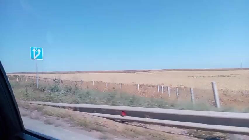 The video shows a view from inside a vehicle traveling along a road. The landscape outside is rural, with a large, flat, dry field stretching towards the horizon under a clear blue sky. A fence runs along the edge of the field. Several signs are visible at different points in the video. The first sign, seen around the 00:01 mark, is a white rectangular sign with red lettering and several pictograms, including ones that appear to be prohibitions against littering and smoking. Another sign, a blue square with a white 'P', indicating parking, is seen later, along with a small building. The road appears to be asphalt with white lane markings. The overall impression is of a journey through a sparsely populated, dry agricultural area.