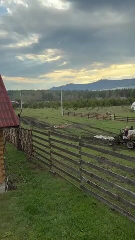 The video shows a small green vehicle with two occupants, likely adults, driving across a grassy field. The vehicle is pulling a small trailer loaded with what appears to be bagged items and what looks like pieces of red material or debris, spread on the ground. The scene is framed by a wooden fence in the foreground, with a portion of a building visible on the left. In the background, there are trees and a distant mountain range beneath a cloudy sky with patches of sunlight. The ground is muddy in places, suggesting recent rain or damp conditions.