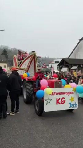 A red tractor with a decorative float is passing through a crowd in a European village. On the float are a group of people dressed in pink and white outfits with crowns. The tractor is decorated with balloons. There is a sign on the front that says "Aktivenwagen KG Jogsch". The scene is outdoors on a cloudy day. There are buildings and trees in the background.