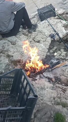 A short video shows two people sitting and standing on a rocky outcropping near a body of water, with fishing rods and crates nearby. A small fire burns on the rocks, and one person appears to be preparing food, possibly meat, stored in a blue bag inside one of the crates. The scene suggests a fishing outing with a small campfire.