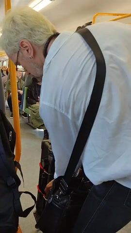 A man with blonde hair and glasses, wearing a white shirt and black pants, is seen from the back as he adjusts luggage on a train. He has a black strap across his shoulder and a black bag attached to his waist. To his left, another black bag is visible. In the background, other passengers are seated on the train. One passenger, wearing a dark hat and green skirt, is looking at their phone. The train's interior is visible with yellow poles and gray seats.