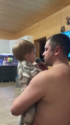 A shirtless man holds a toddler. The man has short dark hair and a silver chain around his neck. The toddler has blonde hair and is wearing pajamas with gray and white cloud patterns. The man holds a small microphone to the toddler's mouth. They appear to be in a wooden cabin. Behind them is a white kitchen and a television mounted on the wall. Decorations hang on the wooden wall.