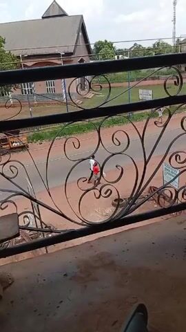 The video shows a view from a balcony overlooking a street. The balcony has an ornate black metal railing with swirling designs. Below, two people, likely children, are walking down the street away from the camera. One is wearing a white shirt and the other a red shirt. A yellow auto-rickshaw passes in the same direction as the children. In the background, across the street, there is a building that appears to be a church, with a grey roof and a cross on top. A grassy field surrounds the church. On the balcony floor is a wooden bench, a flashlight, a solar panel charger, and part of a person's leg in blue jeans. The weather appears to be partly cloudy.
