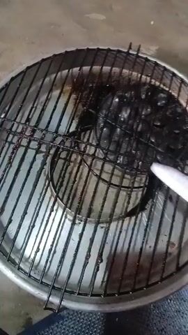 A close-up, overhead shot shows a person's hand holding a knife with a green handle. The knife is being used to scrape a charred object on a metal grill rack that sits inside a shallow metal pan. The object on the grill is difficult to discern due to extensive charring. The background appears to be a concrete floor.
