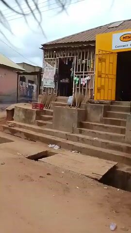 The video shows a street scene with several stalls and buildings lining a dirt road. In the first frame, there is a pink building on the left with posters on its exterior. A drain runs across the foreground. The second frame shows a person in a green and white outfit next to a cart laden with goods. The third frame shows a woman in a yellow top standing in front of a stall. The fourth frame depicts people gathered around a green umbrella, and further down the street, several motorcycles are parked. The overall environment appears to be a marketplace or a busy street in a rural or semi-urban setting. The lighting suggests it is daytime.