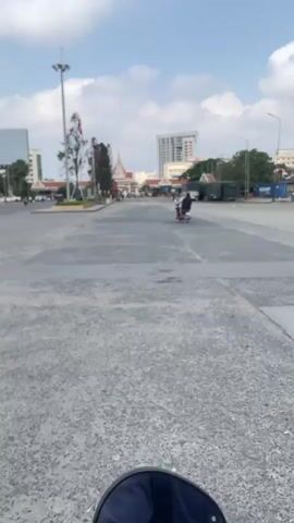 A lone person rides a moped down a wide, paved street under a partially cloudy sky. The street is textured, appearing like a concrete aggregate mix. Buildings, including a prominent high-rise, are visible in the background. A few trees and flagpoles with flags stand along the side of the street. Parked trucks and vehicles are to the right side. The scene appears to be shot from a static position.