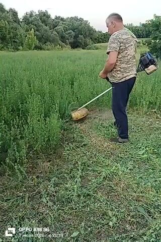 A man in a camouflage t-shirt and dark pants is using a string trimmer to cut tall green grass in a field. He is holding the trimmer with both hands and appears to be moving it back and forth to cut the grass. The field is densely covered with vegetation, and there are trees in the background. The sky is overcast. The video is a screen recording from a phone, indicated by the watermark in the lower left corner which reads "OPPO A98 5G" and the date "12 июля 2023 г. 20:19".