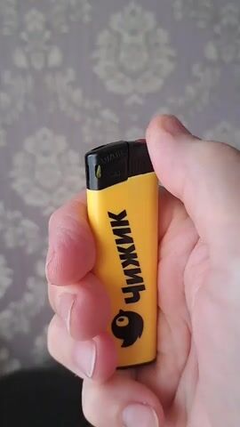 A hand holds a yellow disposable lighter with a black cap. The lighter has a cartoon bird graphic and the Russian word "ЧИЖИК" (Chizhik) printed on it. The hand operates the lighter, and a flame ignites and burns steadily. The background is a wall with a gray floral pattern. The lighting suggests an indoor setting, possibly during daylight. The action is simple: the user is lighting the lighter. No specific time of day, weather, or emotional cues are discernible from the image. The location context provided is Lytkarino, Russia.