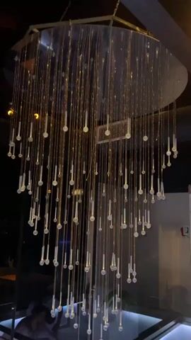 This is a video of a modern chandelier made of thin, golden strands with small, clear cylindrical or capsule-shaped objects hanging from them at varying lengths. The chandelier is suspended from a ceiling, which appears to be dark wood. The light from the chandelier is warm and golden, and it illuminates the surrounding area, casting subtle shadows. In the background, there are glimpses of a dark interior with some out-of-focus lights and structures. At the very bottom of the frame, a blurry reflection of a person or people can be seen on what appears to be a reflective surface. The video is shot from a low angle looking up at the chandelier.