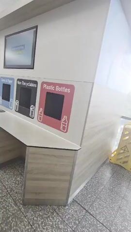 The video opens with a close-up of an airport recycling station with multiple bins labeled for different types of waste. The bins are labeled "Plastic Bottles", "Non Recyclables", "Cans & Paper", "Plastic Bottles", "Food", and "Glass". A screen above the bins displays a message that reads "Keep everything in your bag". The camera then pans left, revealing more of the airport terminal. A young man wearing a t-shirt and shorts walks away from the camera and towards the departure gates. Other travelers are seen in the background, waiting to pass through security. The environment appears to be clean and modern, with bright lighting and a tiled floor. The overall impression is that of a busy airport environment, with travelers preparing for their flights.