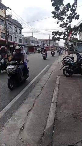 The video shows a street scene with several motorcycles driving on a paved road. On the left side of the road, there are multi-story buildings. On the right side of the road, there is a sidewalk with parked motorcycles. The sky is cloudy.