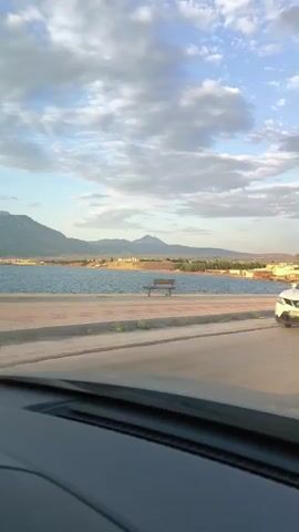 The video captures a view from inside a vehicle, looking out at a roadside scene. In the foreground, the dashboard of the vehicle is visible. The road runs parallel to a body of water, likely a lake or sea, with a distant shoreline featuring hills and mountains. Several cars are driving on the road. One white SUV passes by, and a black SUV follows shortly after. Later, a silver SUV is seen, with a person visible inside. The sky is partly cloudy, with soft lighting suggesting it might be late afternoon or early evening. The overall impression is of a scenic drive along a coastal or lakeside route.