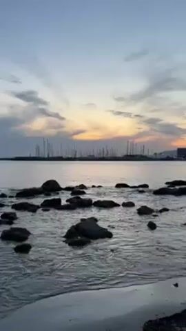 The video shows a seascape with a sunset sky. The water is calm with small waves lapping against rocks in the foreground. In the distance, there are many sailboat masts visible in a harbor or marina. The sky is a mix of blues, greys, and oranges from the setting sun. There is a small strip of sandy beach in the very foreground.